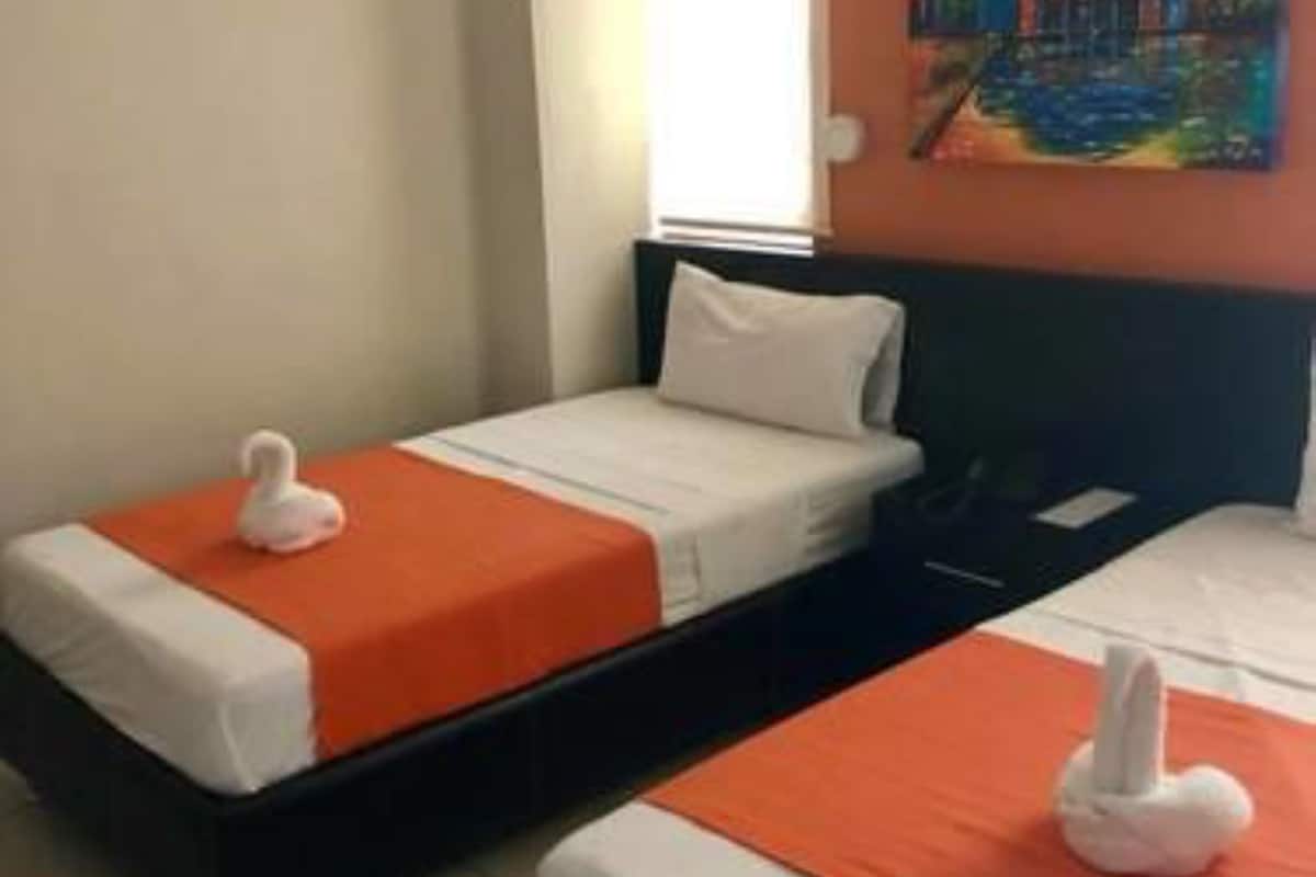 A hotel room with two beds, each with white bedding and orange headboards. There is a painting on the wall above the beds, and an air conditioning unit mounted on the wall.