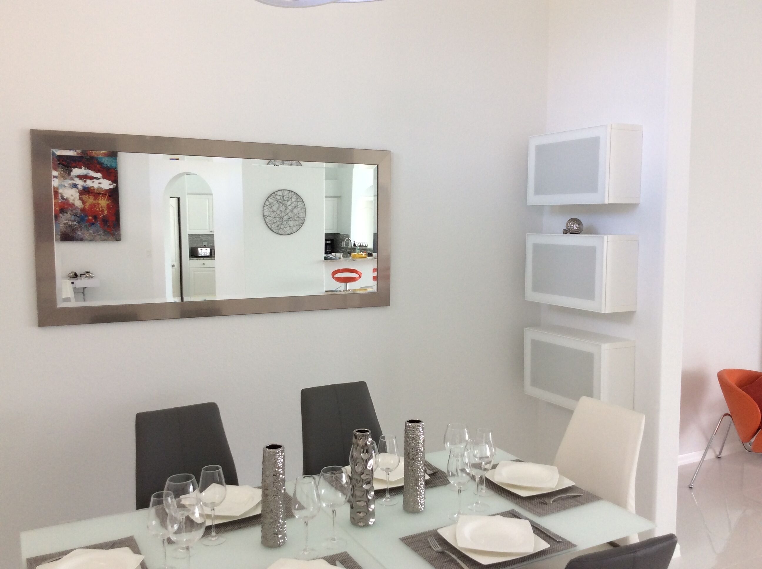 A dining area with a table set for four, a large mirror, and a modern kitchen visible in the reflection.