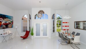 A spacious, modern dining area with a high ceiling, large windows, and a central chandelier.