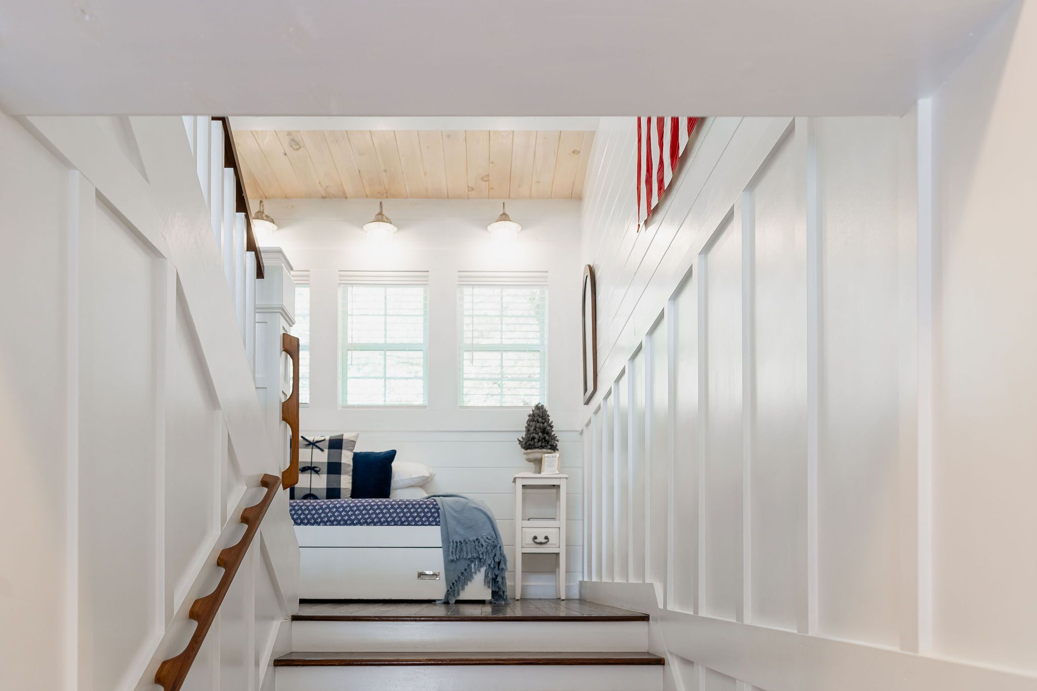 Un couloir étroit mène à une chambre dotée d’un escalier en bois, de murs blancs et d’une petite table de chevet sur laquelle est posé un objet décoratif.