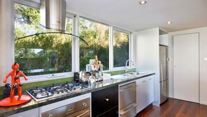 Une cuisine moderne dotée d’une cuisinière à gaz, d’appareils électroménagers en acier inoxydable, d’une grande fenêtre offrant une vue sur les arbres et d’un design minimaliste.