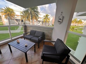 A balcony with a sofa and a table, a stone wall, and a view of palm trees and buildings.