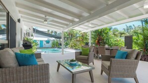 A covered outdoor seating area with wicker furniture, a glass coffee table, and a view of a swimming pool and lush greenery. - Paradise on Ruby - Port Douglas (Port Douglas)