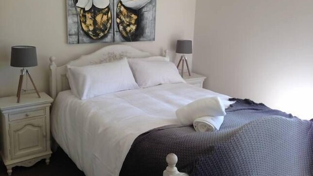 A neatly made bed with white linens, two bedside lamps, and a large floral painting above the bed. - Fairway Views Accomadation (Warragul)
