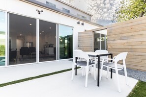 Ein modernes Haus mit einer Terrassenfläche, die einen Tisch und Stühle beinhaltet, großen Schiebeglastüren, die zu einem Innenraum führen, und einem Holzzaun.
