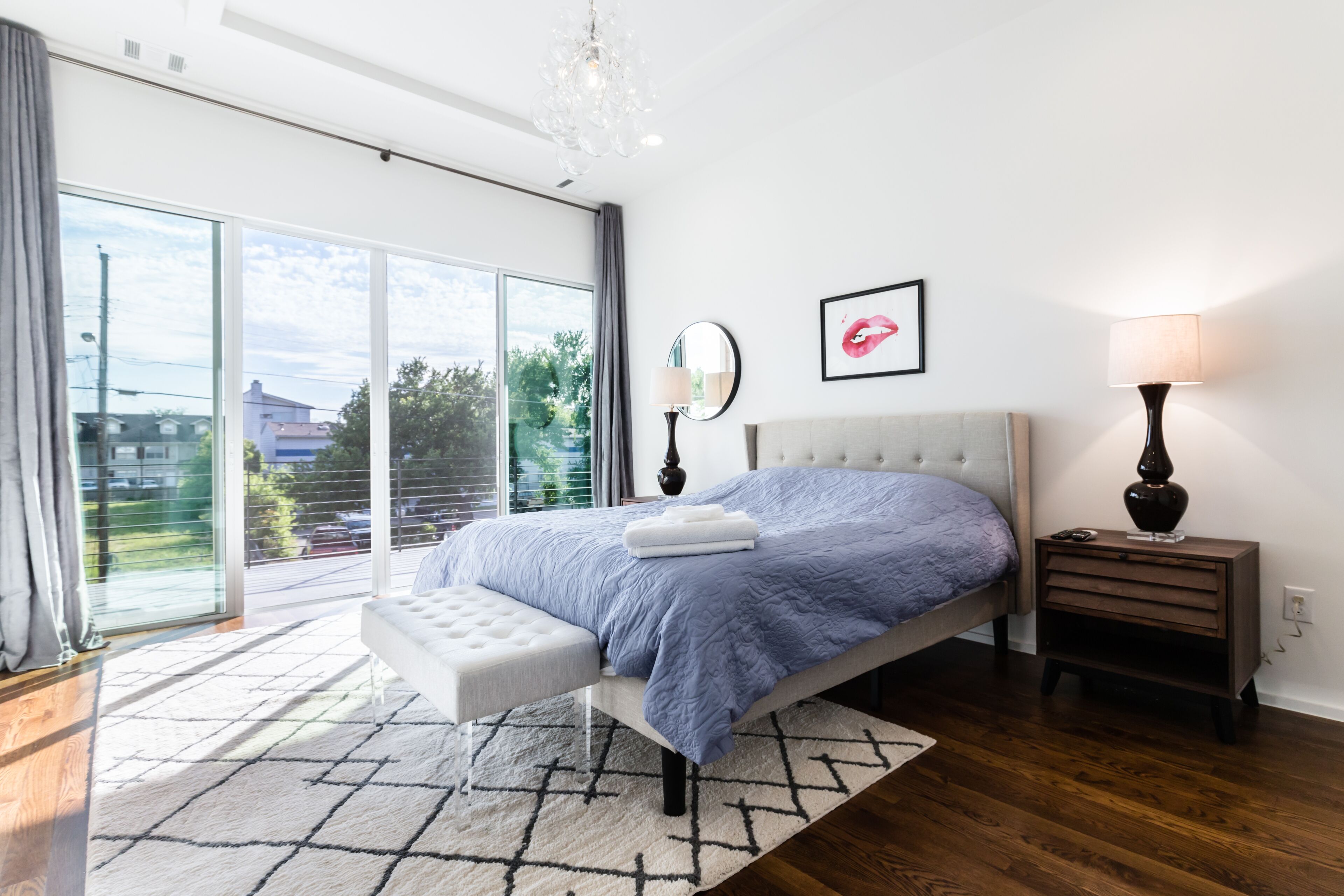 A modern bedroom with a large bed, a bedside table, a nightstand, a chandelier, and a view of the outdoors.