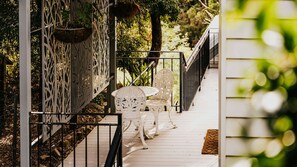 Une terrasse en bois, avec des chaises et une table en métal peintes en blanc, entourée d’une clôture en métal et de verdure.