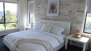 Une chambre avec un grand lit, une tête de lit, une table de chevet, une œuvre d’art encadrée et une vue sur l’extérieur.