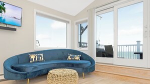 A modern living room with a curved blue sofa, two patterned cushions, a woven ottoman, and a flat-screen TV on the wall.