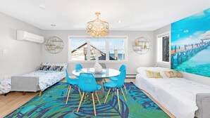 A modern living room with a round table, blue chairs, a white sofa, and a large window with a view of a pier.
