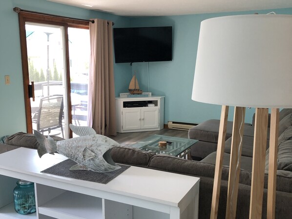 A living room with a sofa, a coffee table, a TV mounted on the wall, and a sliding glass door leading to an outdoor area. - Year round beach house 5 minute walk to beach with all the comforts of home. (York)