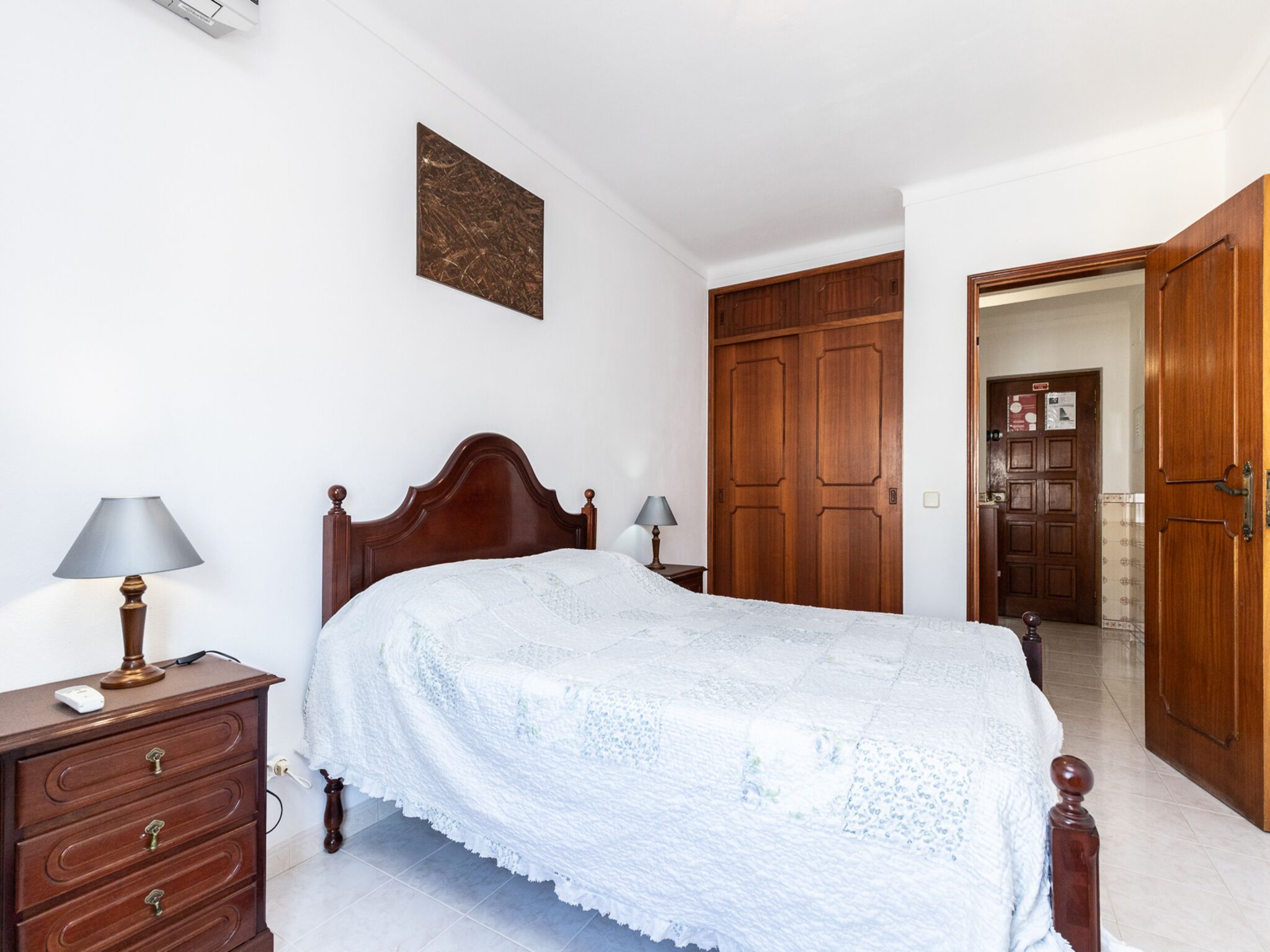Een slaapkamer met een houten bed, nachtkastje met lamp, een dressoir en een kledingkast.