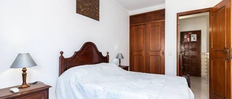 Een slaapkamer met een houten bed, nachtkastje met lamp, een dressoir en een kledingkast.