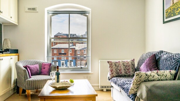 A cozy living room with a large window, a striped armchair, a sofa with patterned cushions, and a wooden coffee table. - 30 Woodsmill Quay - sleeps 3 guests  in 1 bedroom (York)