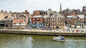 A riverside promenade with historic buildings, a boat docked on the water, and a church spire in the background. - 30 Woodsmill Quay - sleeps 3 guests  in 1 bedroom (York)