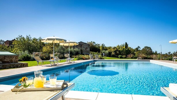 Een zwembad met ligbedden en parasols, omgeven door groen en een stenen gebouw.