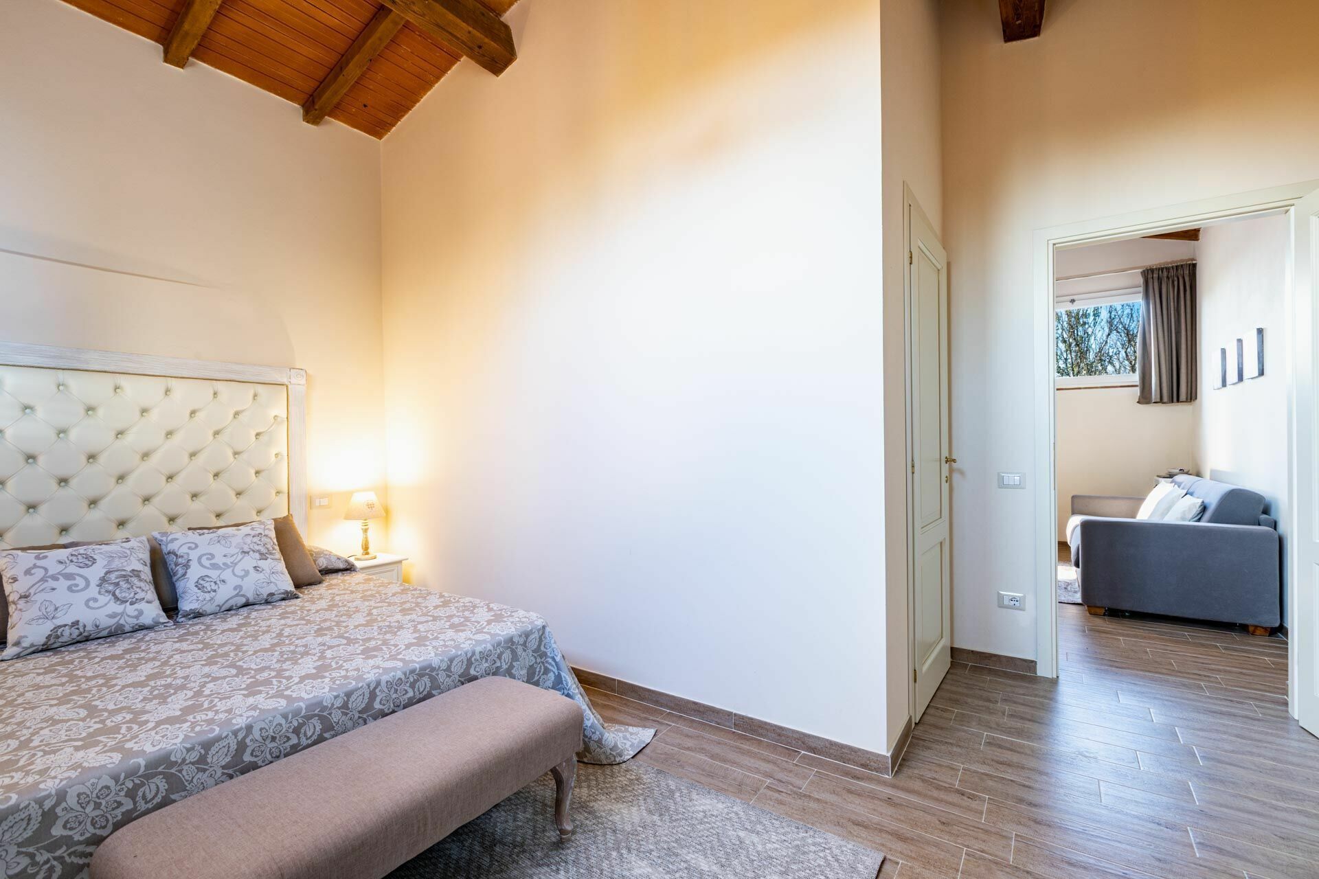 Un dormitori amb llit, taula de nit, làmpada i vista a una altra habitació.