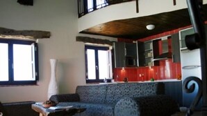 A living room with a sofa, a coffee table, and a window. There is a kitchen area with red cabinets and a countertop.