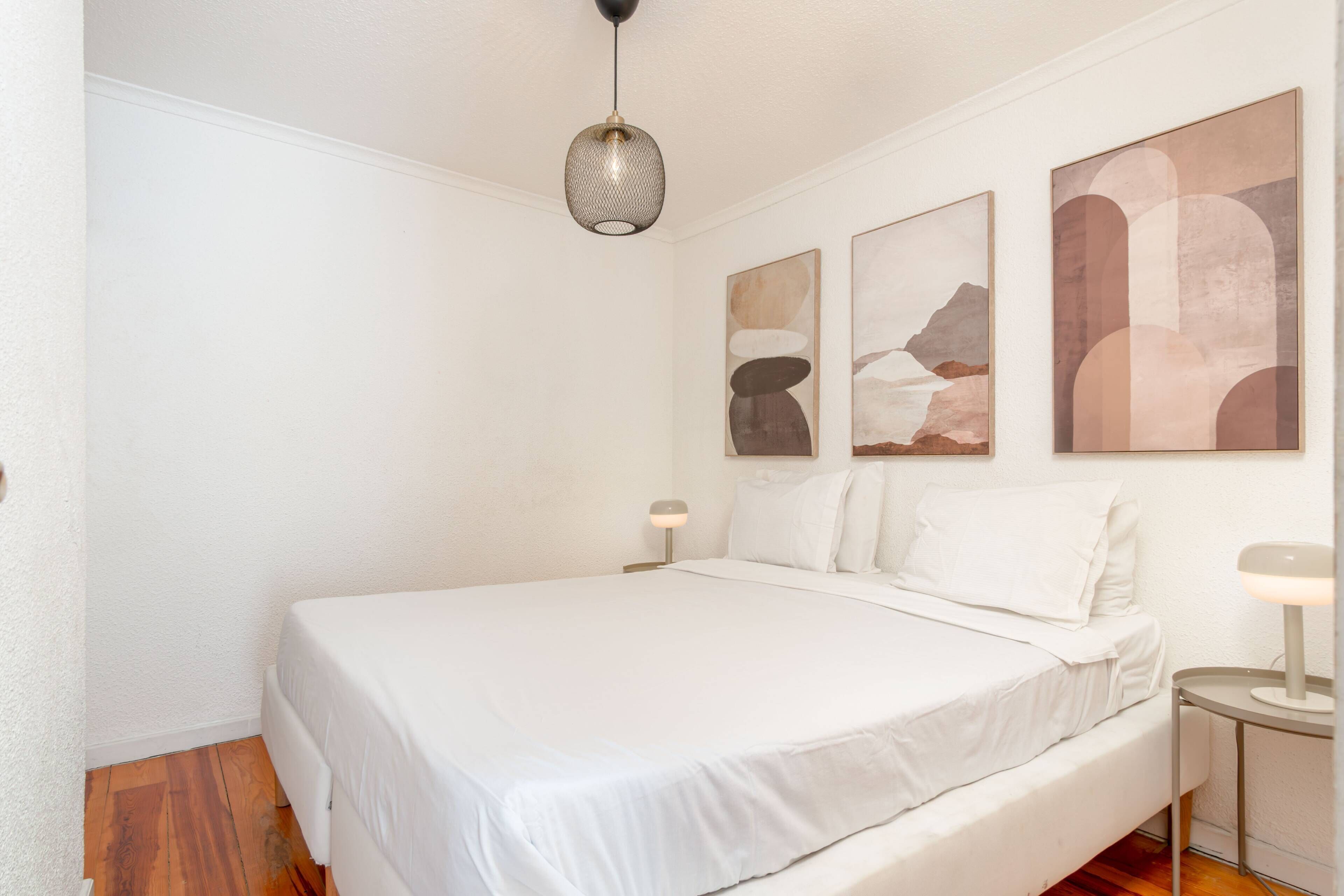 A bedroom with a large bed, two bedside tables, a hanging light fixture, and three abstract wall art pieces.