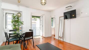 A room with a dining table, chairs, a sofa, a TV mounted on the wall, and a potted plant.