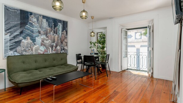 A living room with a green sofa, a black coffee table, and a cityscape painting on the wall. There are three pendant lights, a dining area with a table and chairs, and a balcony with a railing. - Typical Duplex Apartment in Bairro Alto (Lisboa)