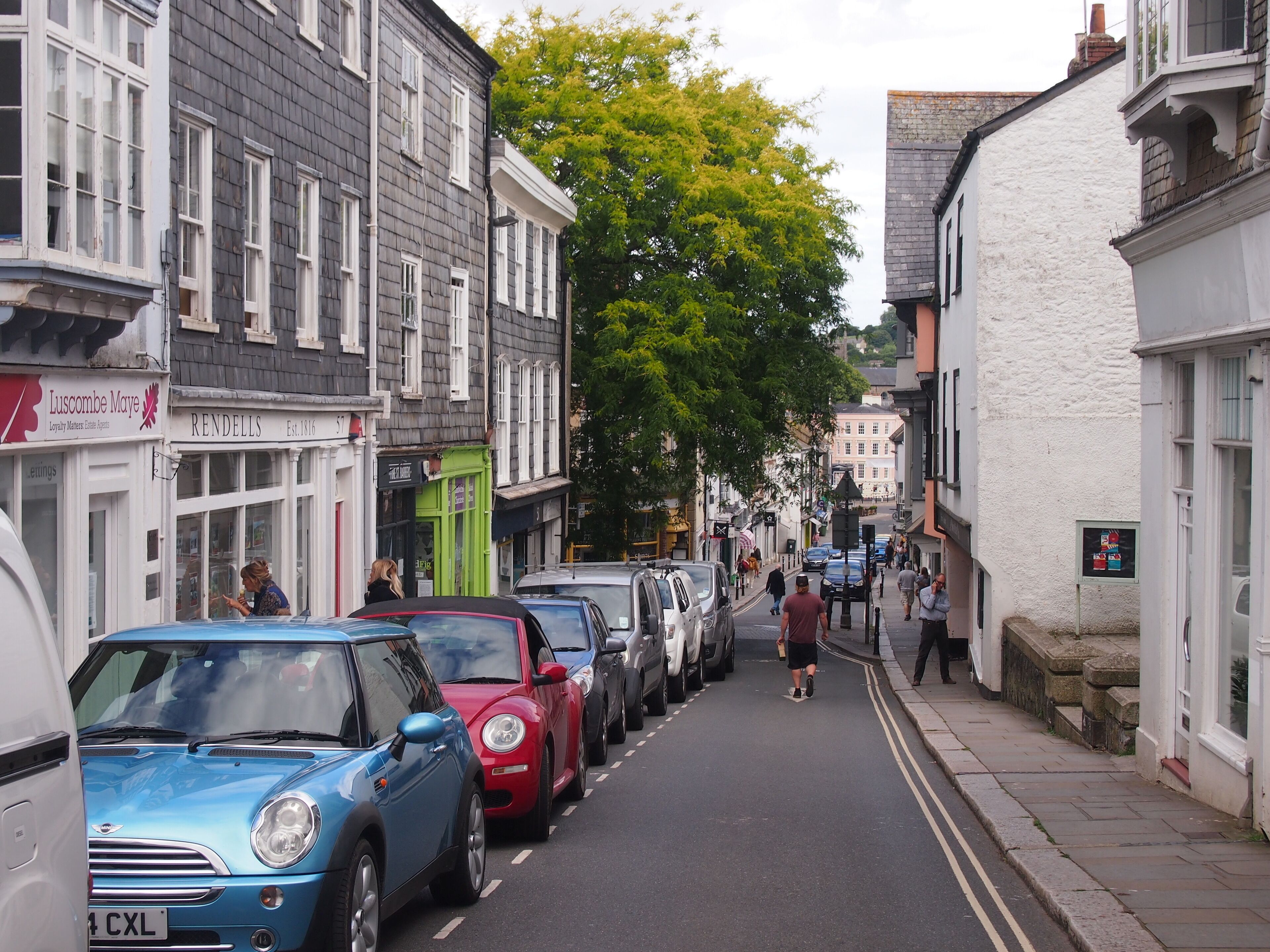 A narrow street lined with historic buildings, parked cars, and pedestrians.