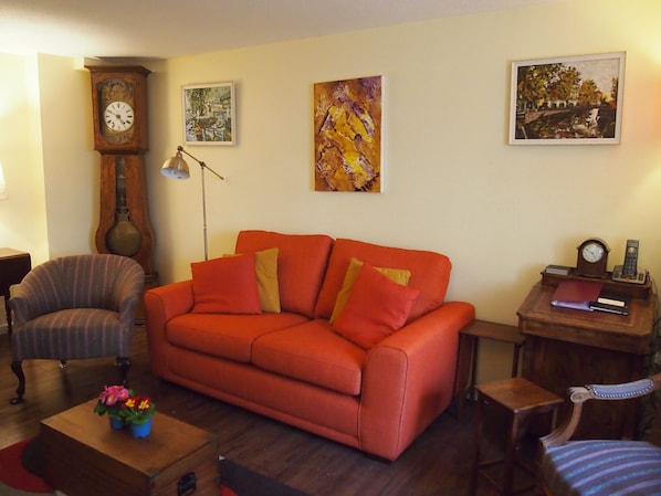 A living room with a red sofa, a wooden side table, a floor lamp, a clock, and two framed paintings on the wall.