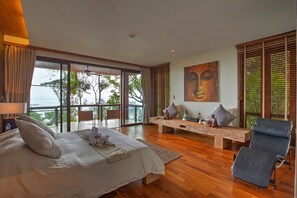 Une chambre spacieuse avec un grand lit, un coin salon avec un banc et une vue sur l’océan grâce à de grandes fenêtres.
