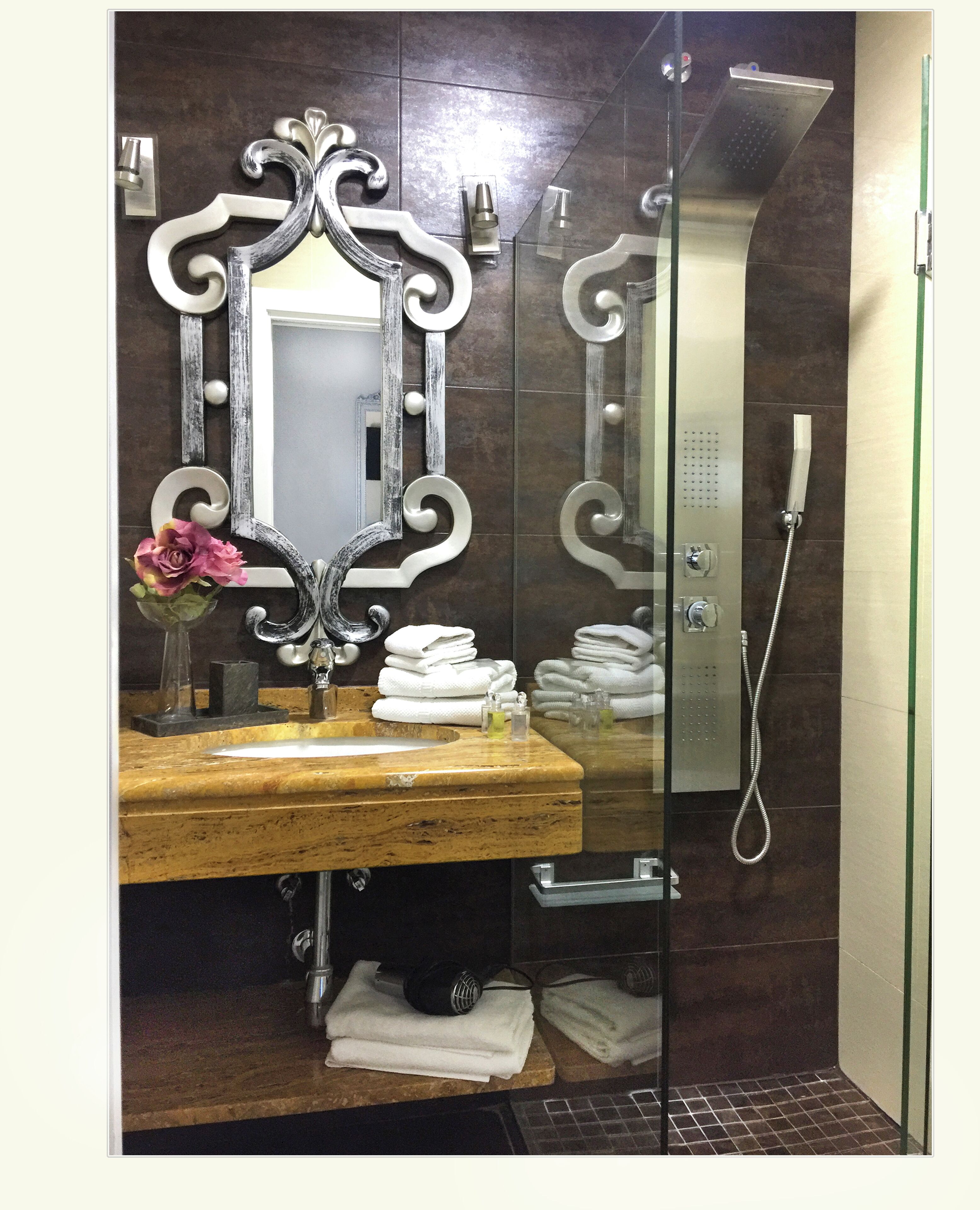Ein Badezimmer mit einem Holzkosmetikschrank, einem großen Spiegel mit aufwendigen Verzierungen, einer Dusche mit Duschbrause und ordentlich gestapelten Handtüchern.