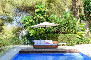 Un área junto a la piscina con cubierta de madera, un sofá blanco con cojines y una sombrilla grande.