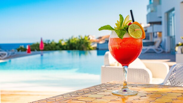 A cocktail with garnishes on a table by a poolside, with a clear view of the pool and a building in the background.