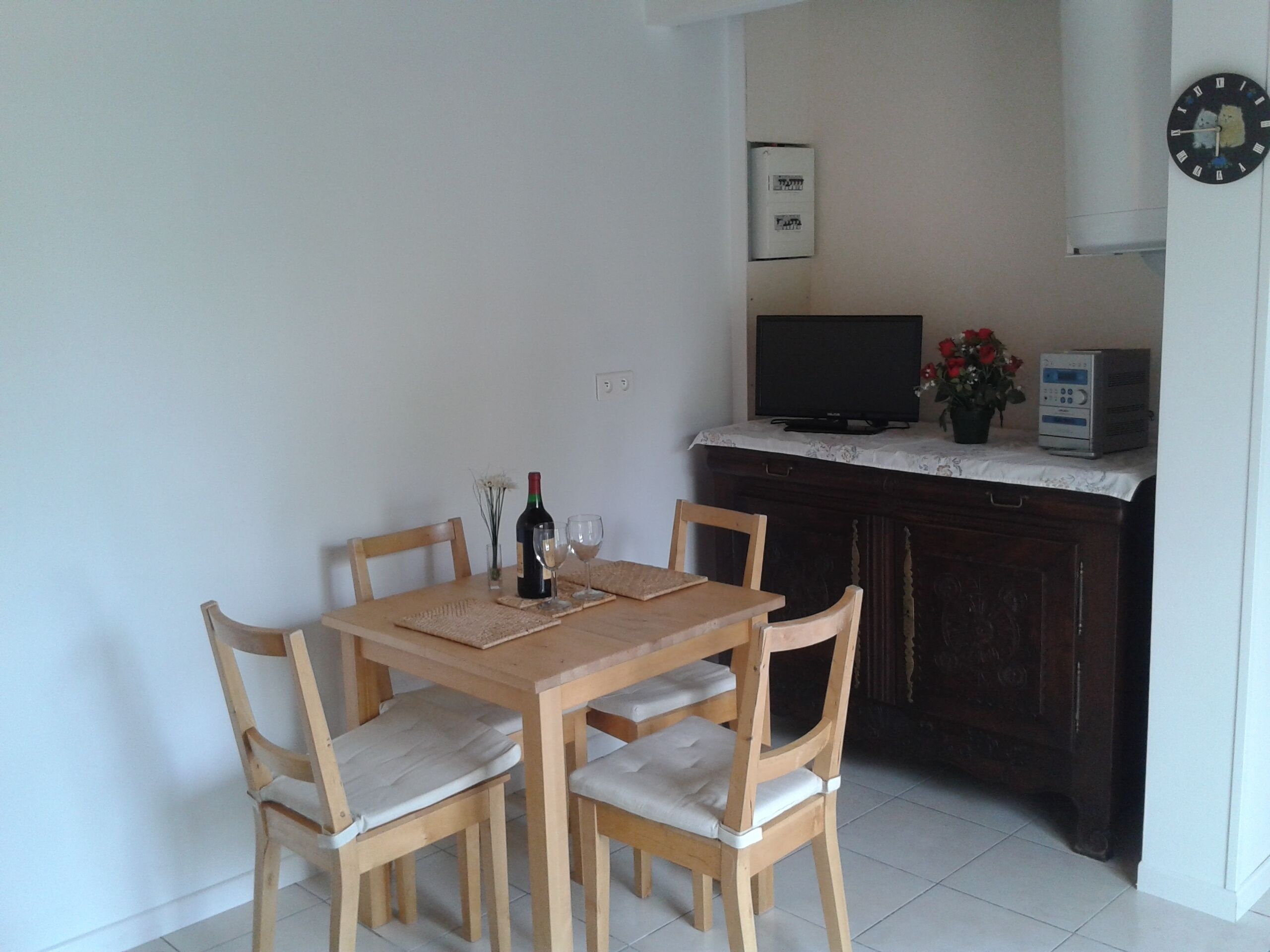 A dining area with a wooden table and chairs, a bottle of wine, and two wine glasses. A television and a small appliance are on a counter to the right.