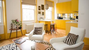 Un salon moderne avec un canapé et des fauteuils, une table basse et une petite table d’appoint sur laquelle se trouve un vase. On y trouve une fenêtre avec des stores, un radiateur et un coin cuisine avec des meubles jaunes et un tabouret de bar.