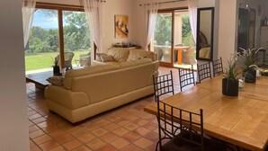 A spacious living room with a large sofa, a wooden dining table, and a view of a lush garden through large windows. - Modern villa - 5 bedrooms, in Uchaux village - private pool & amazing views (Uchaux)