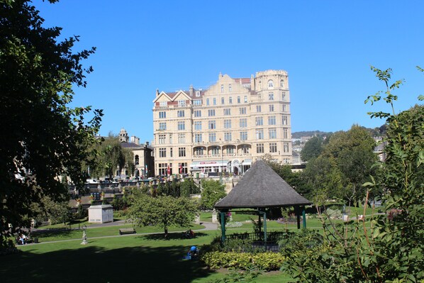 A large, historic building with multiple stories and a prominent tower, surrounded by lush greenery and a park area with a gazebo.