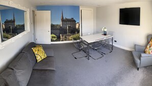 Un salon moderne avec un canapé gris, une table à manger et des chaises, un grand miroir et une fresque représentant un paysage urbain.