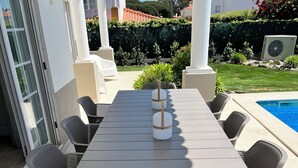 Una terrazza esterna con un tavolo rettangolare e sei sedie, circondata da una piscina e vegetazione.