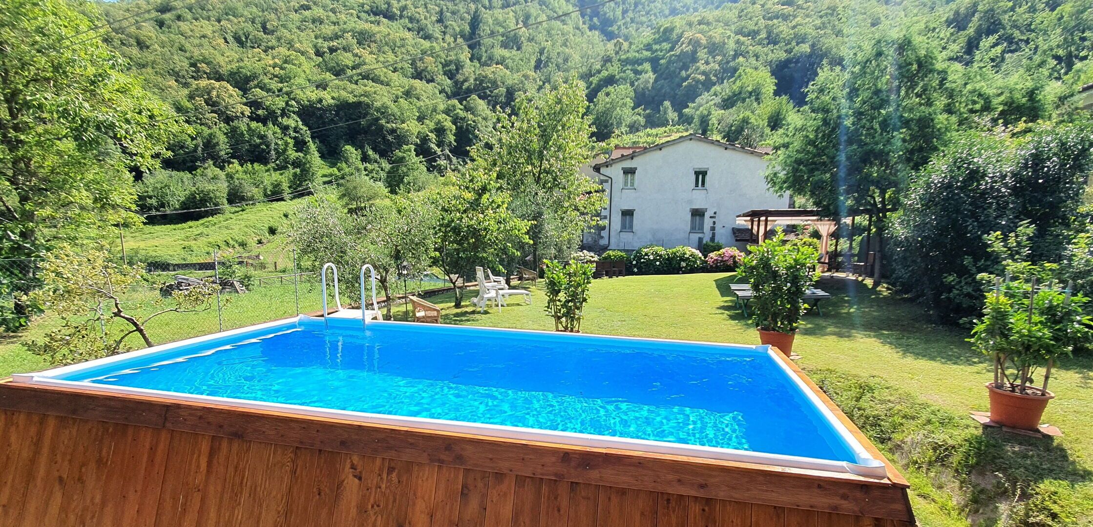 A rectangular swimming pool with a wooden deck, surrounded by a lush green landscape and a white house in the background.