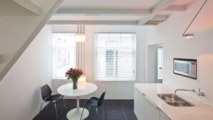 A modern kitchen with a white countertop, a stainless steel sink, and a built-in microwave. There is a dining area with a white table and two black chairs, and a vase with flowers on the table.