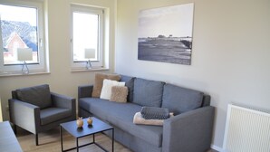 A modern living room with a grey sofa, a small table, and two armchairs. There is a window with a view of a house and a beach scene on the wall.