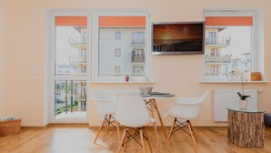 Een moderne eethoek met een tafel en stoelen, grote ramen met oranje jaloezieën, uitzicht op een gebouw en een ingelijst schilderij aan de muur.