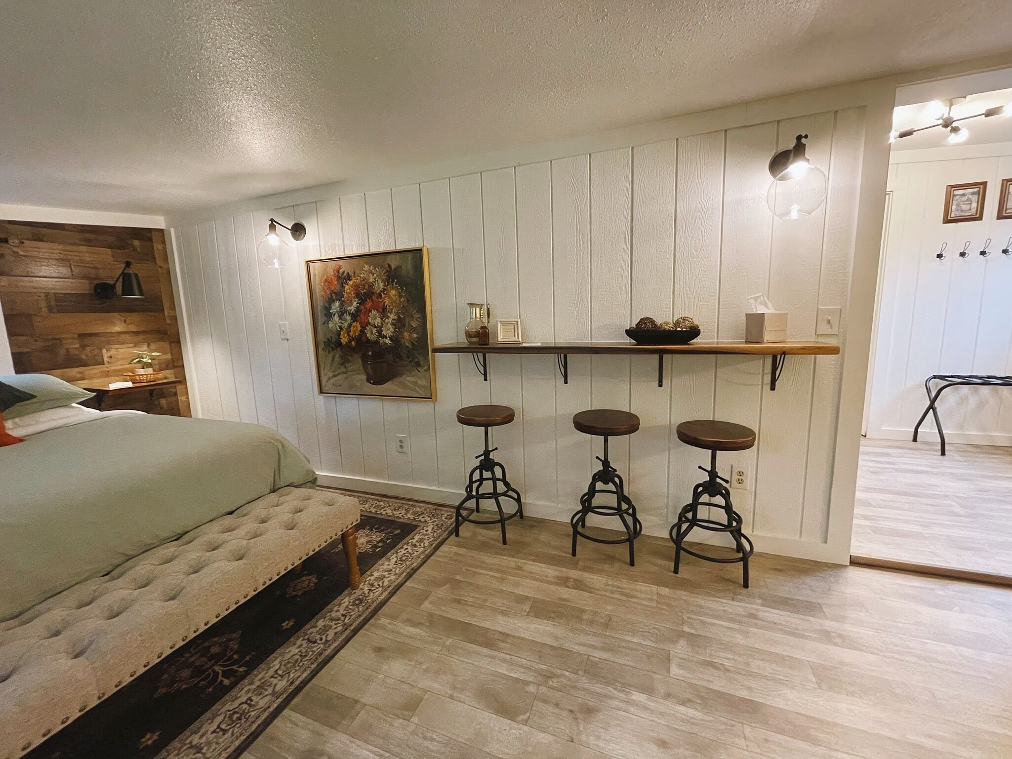 A bedroom with a bed, a nightstand, a lamp, a painting, a shelf with decorative items, and three stools.