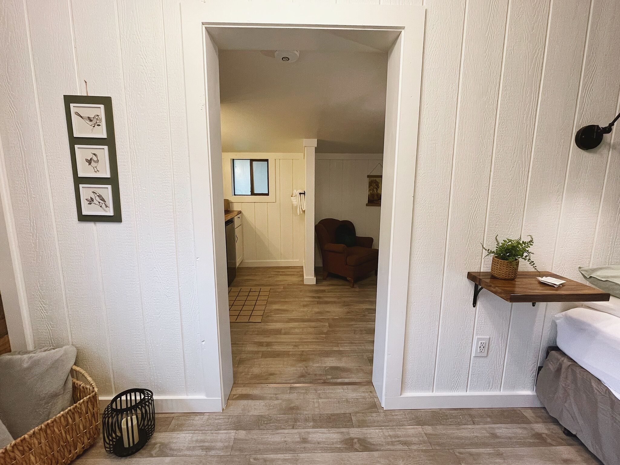 A narrow hallway leading to a room with a bed, a small table with a plant, and a chair. There are framed pictures on the wall and a basket on the floor.
