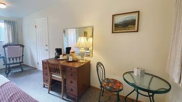 Habitación de hotel con un escritorio de madera, una silla, una mesita con tapa de vidrio, un espejo y un cuadro en la pared.