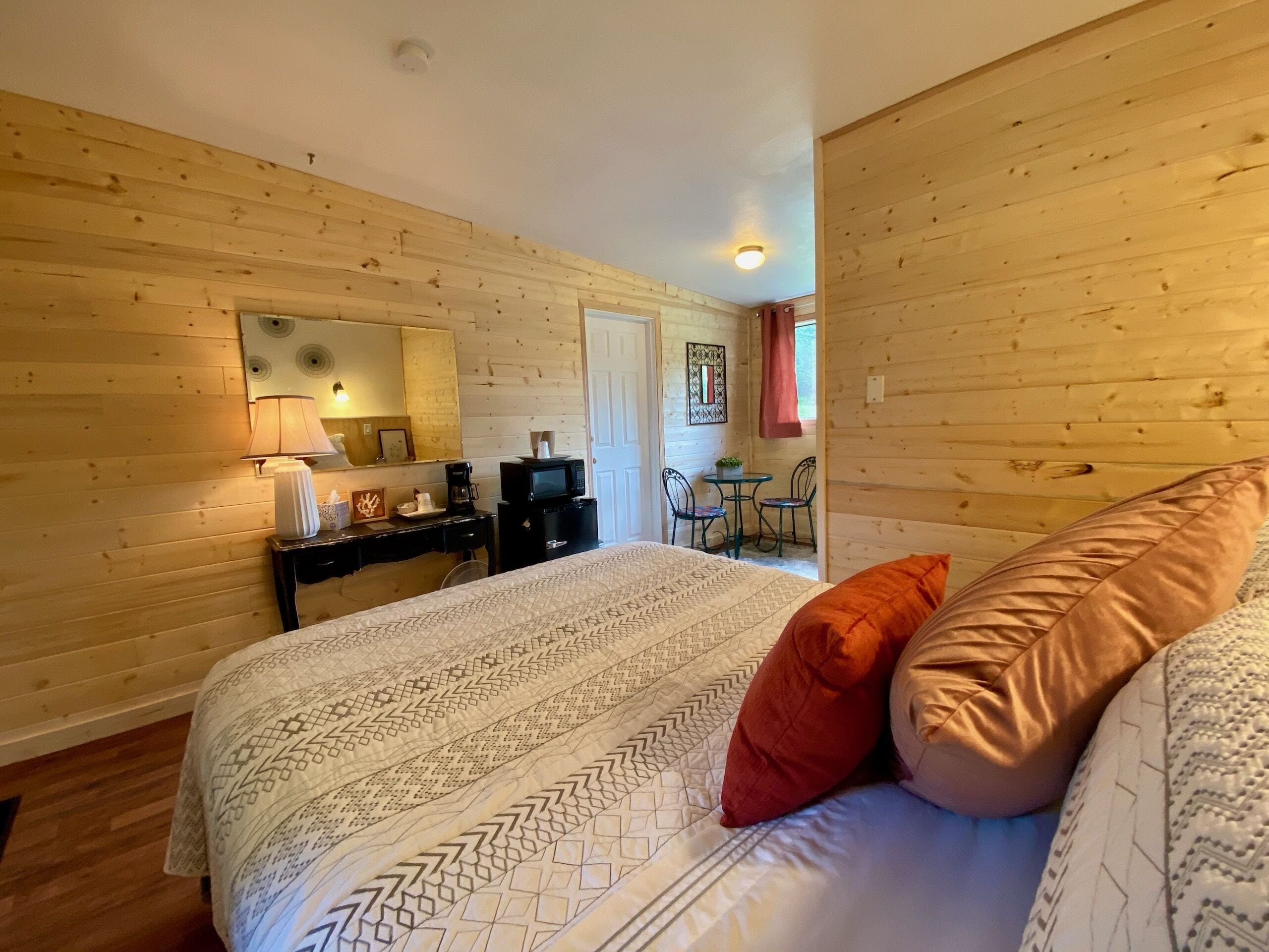 Quarto com cama, almofadas, escrivaninha com abajur, espelho e mesa pequena com cadeiras.