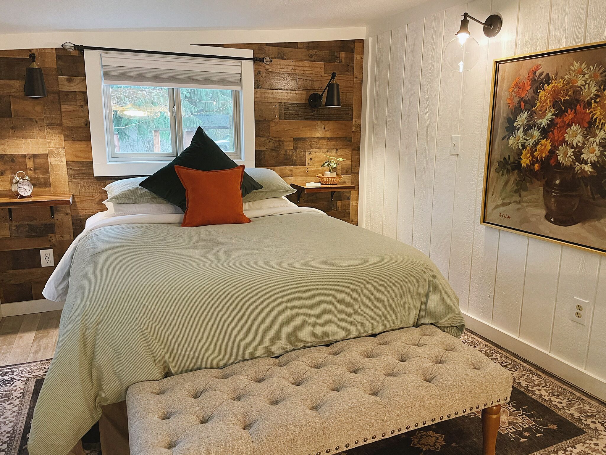 A bedroom with a bed, a bench, a window, a shelf with a plant, and a painting.