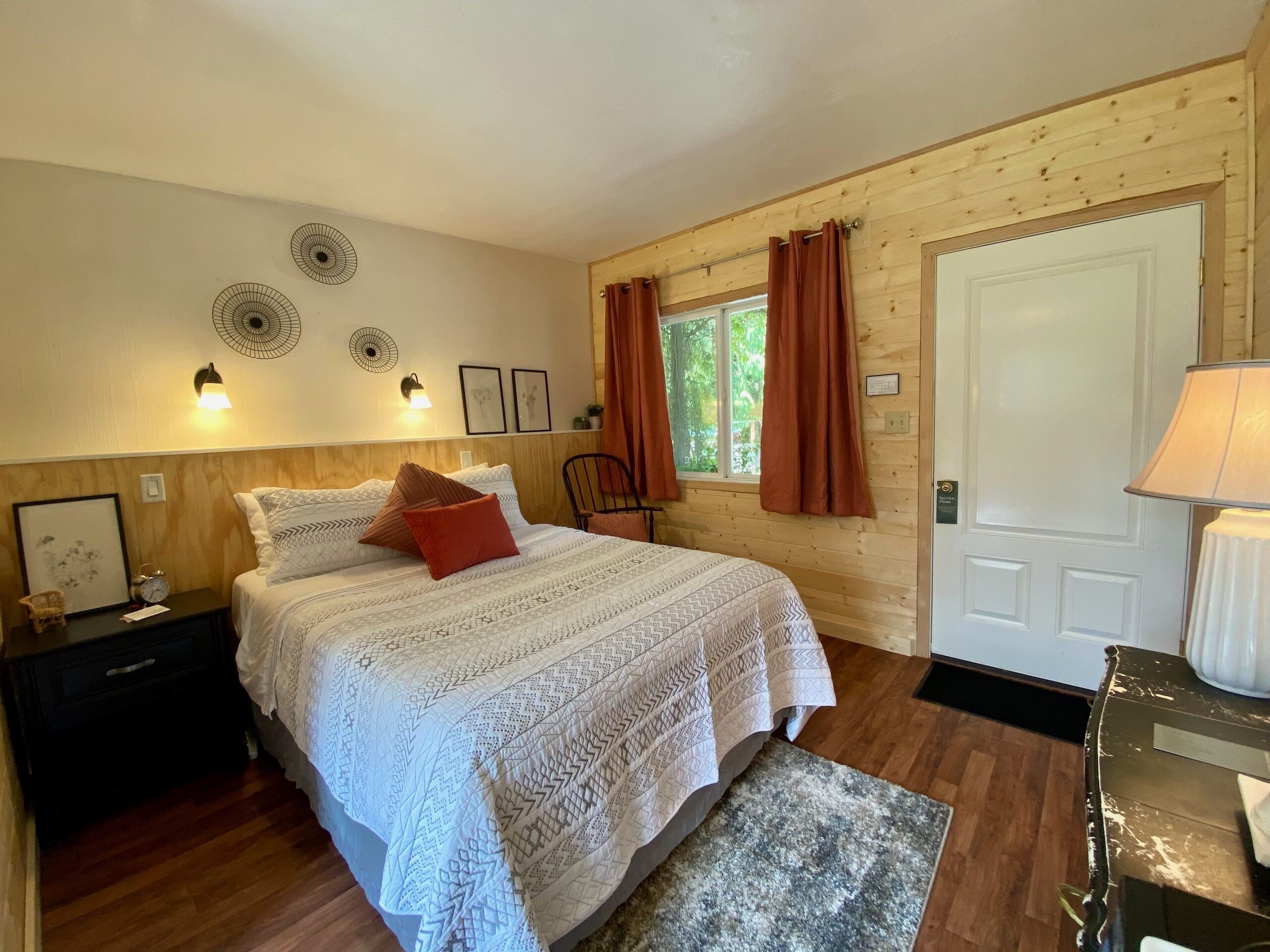 A cozy bedroom with a large bed, wooden walls, a window with curtains, a bedside table with a lamp, and a small chair.