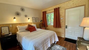 A cozy bedroom with a large bed, wooden walls, a window with curtains, a bedside table with a lamp, and a small chair.
