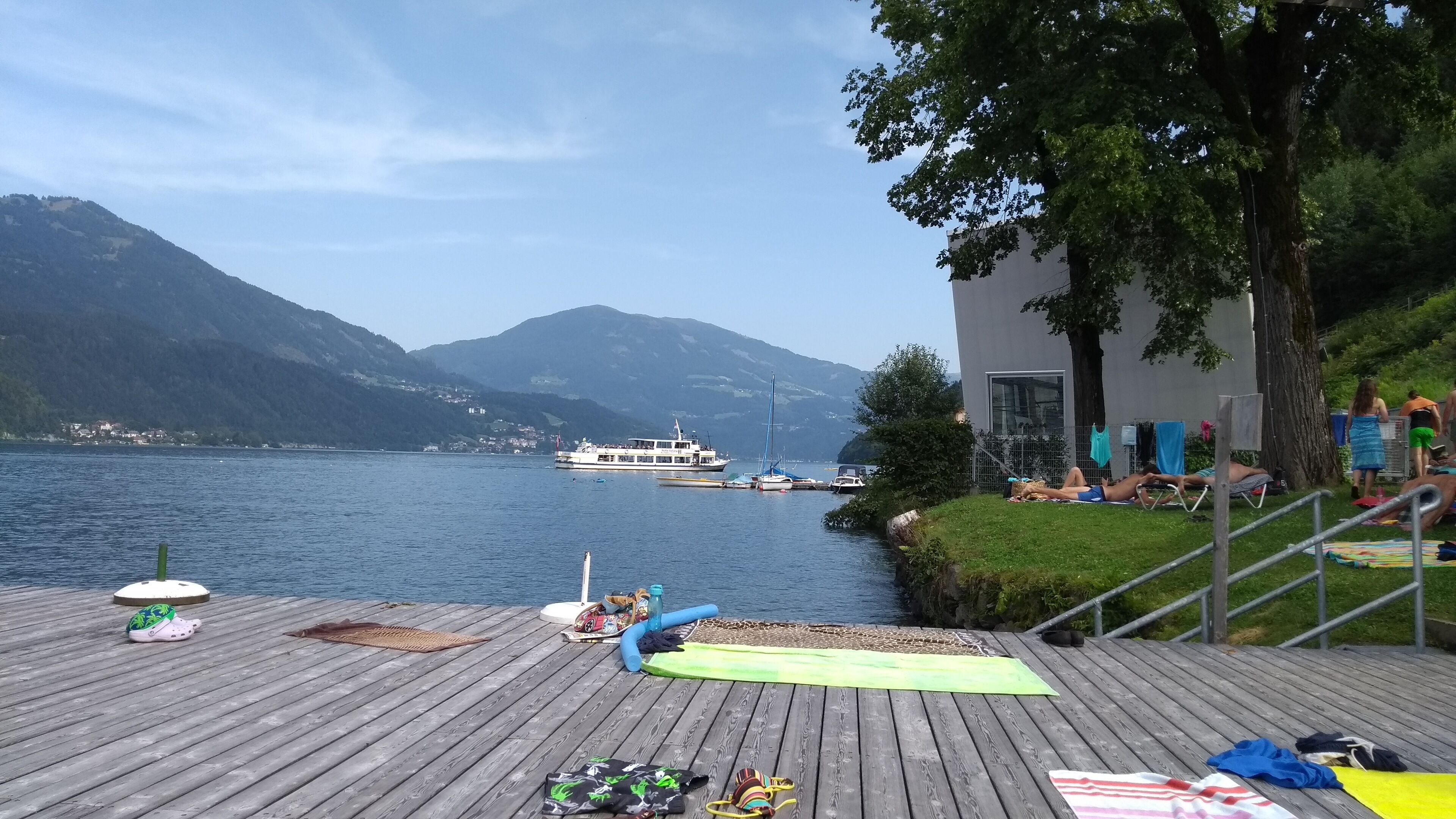 A wooden deck with a view of a lake and mountains, a boat in the distance, and people relaxing on the deck.