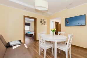 A modern living room with a round dining table, white chairs, a sofa with a patterned cushion, a wall clock, and a flat-screen TV mounted on the wall.